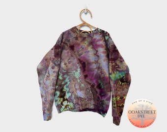 Pode incluir: Uma camisola tie-dye em tons de roxo, castanho, verde e azul. A camisola tem um decote redondo e mangas compridas. Está pendurada num cabide de madeira. A etiqueta na camisola diz "Oak Street Dye, One of a Kind, Made on Oak Street."
