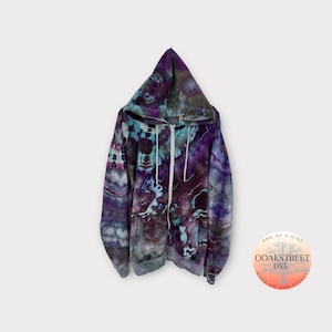 Może przedstawiać: Szara bluza z kapturem w odcieniach fioletu i błękitu z efektem tie-dye i zamkiem błyskawicznym. Bluza ma logo "OOAKSTREET DYE" w prawym dolnym rogu. Wokół logo widnieje napis "ONE OF A KIND" i "MADE ON OAK STREET".