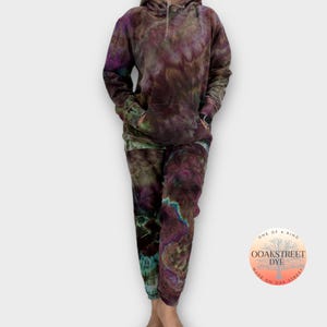 May include: A tie-dye hoodie and sweatpants set in shades of purple, green, and brown. The set features a large pocket on the front of the hoodie and a drawstring waistband on the sweatpants. The clothing is labeled "OOAKSTREET DYE" and "ONE OF A KIND".