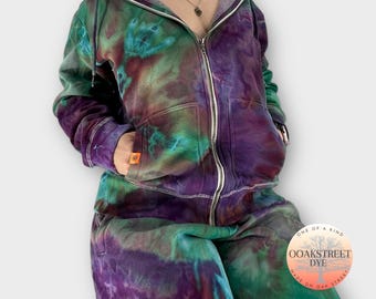 May include: A tie-dye hooded sweatshirt and sweatpants set in shades of purple, green, and blue. The set features a full-zip hoodie with a kangaroo pocket and a drawstring hood. The sweatpants have an elastic waistband and a relaxed fit. The clothing is labelled "OOAKSTREET DYE" and "ONE OF A KIND".