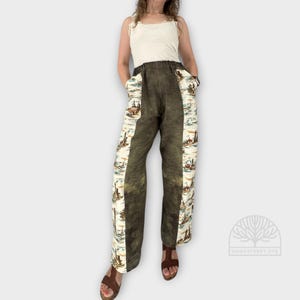 May include: High-waisted pants featuring a solid dark olive green center panel and side panels with a cream background and a multi-colored landscape print. The pants have a wide-leg silhouette and are paired with a sleeveless cream-colored top.