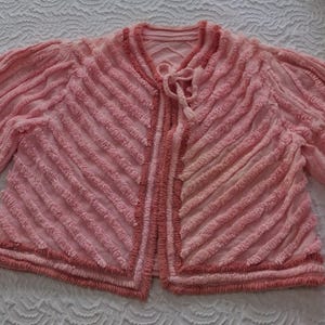 May include: A light pink cardigan featuring a textured diagonal stripe pattern across the front and sleeves. The garment has three-quarter length sleeves, a round neckline, and a front tie closure with matching pink fabric strings at the collar.