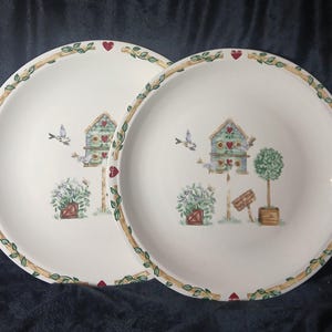 May include: Two white ceramic plates with a decorative birdhouse and floral design. The plates feature a border of green leaves, red hearts, and a light brown trim. The birdhouse is blue, green, and red, with a small bird flying nearby.