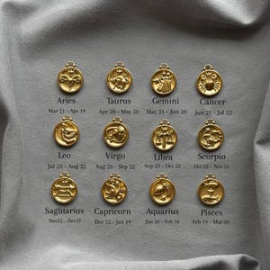 Może przedstawiać: Złote zawieszki z symbolami zodiaku, każda z innym symbolem astrologicznym i odpowiadającymi datami. Zawieszki są ułożone na szarym tle z tkaniny. Tekst zawiera nazwy znak&oacute;w zodiaku i ich daty.
