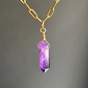 Peut inclure: Un collier en chaîne dorée avec un pendentif en cristal d'améthyste violet. Le cristal est facetté et a une pointe pointue.