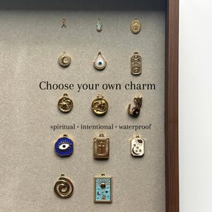 May include: A selection of gold-coloured charms with diverse designs, such as a crescent moon, an eye, and a cross. The text "Choose your own charm" is visible, alongside the words "spiritual, intentional, waterproof."