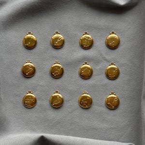 Może przedstawiać: Dwanaście złotych zawieszek zodiakalnych z pętelką u g&oacute;ry do przymocowania do łańcuszka. Każda zawieszka przedstawia inny wz&oacute;r konstelacji. Zawieszki ułożone są na szarej powierzchni materiału.