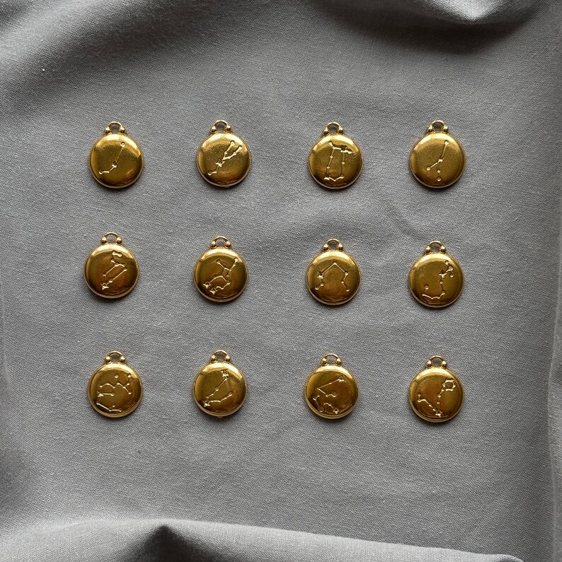 Może przedstawiać: Dwanaście złotych zawieszek zodiakalnych z pętelką u g&oacute;ry do przymocowania do łańcuszka. Każda zawieszka przedstawia inny wz&oacute;r konstelacji. Zawieszki ułożone są na szarej powierzchni materiału.