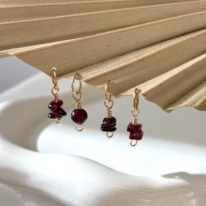 May include: Four gold-toned metal charms featuring deep red garnet stones. Each charm is suspended from a small circular jump ring. The stones vary in shape, including irregular chip-like pieces, a smooth round bead, and small rectangular segments.