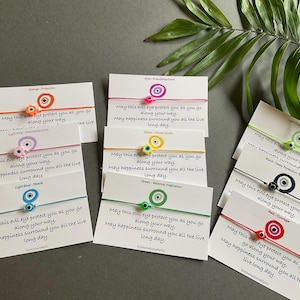 Könnte beinhalten: Eine Sammlung von Evil-Eye-Armbändern auf Karten, jedes mit einer unterschiedlich farbigen Kordel und Augenperle. Die Karten tragen den Text "May this evil eye protect you as you go along your way. May happiness surround you all the live long day."