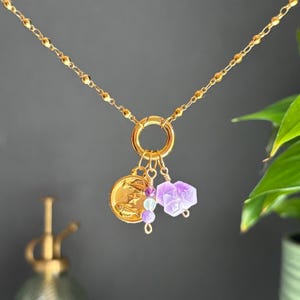 May include: A gold-colored necklace with a delicate chain and a large circular clasp. The necklace features a gold coin charm with a celestial design, along with purple and blue beads, and a faceted amethyst crystal pendant. The necklace is a piece of jewelry.
