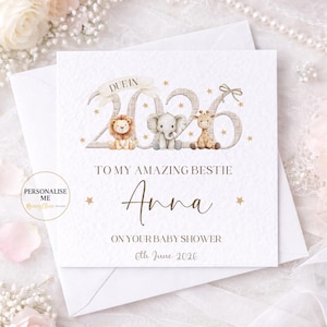 Peut inclure: Carte de baby shower blanche avec le texte "TO MY AMAZING BESTIE Anna ON YOUR BABY SHOWER 6th June 2026". La carte présente des illustrations d'un lion, d'un éléphant et d'une girafe, avec l'année "2026" en arrière-plan.