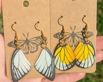May include: Two pairs of butterfly earrings on brown card stock. One pair features white butterfly wings with black markings, and the other pair features yellow butterfly wings with black and orange markings.