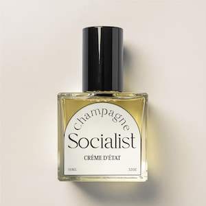 May include: A square glass perfume bottle with a black cap. The bottle contains a light yellow liquid and has a white label with the words "Champagne Socialist" and "Crème d'État". The bottle is labeled 10 ml and .32oz.