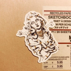 Op de afbeelding: Een sticker met een gedetailleerde illustratie van Medusa met slangen als haar, in bruin- en goudtinten. De sticker ligt op een bruin papieren oppervlak naast een schetsboek.