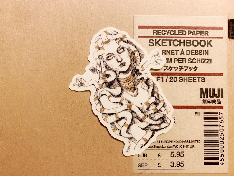 Op de afbeelding: Een sticker met een gedetailleerde illustratie van Medusa met slangen als haar, in bruin- en goudtinten. De sticker ligt op een bruin papieren oppervlak naast een schetsboek.