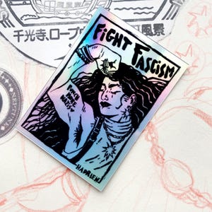 Puede incluir: Pegatina hologr&aacute;fica con una ilustraci&oacute;n en blanco y negro de una persona con pelo largo, un pu&ntilde;o en alto y las palabras "FIGHT FASCISM". La pegatina tambi&eacute;n presenta el texto "PUNCH NAZIS" en el brazo.