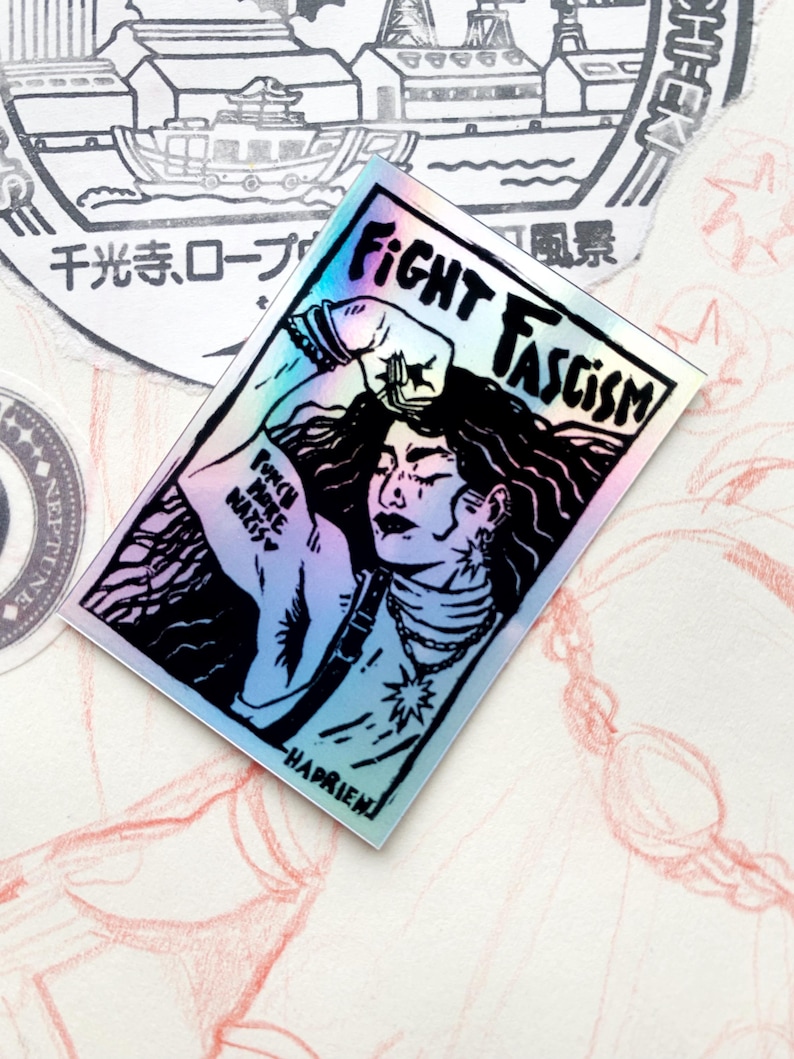 Puede incluir: Pegatina hologr&aacute;fica con una ilustraci&oacute;n en blanco y negro de una persona con pelo largo, un pu&ntilde;o en alto y las palabras "FIGHT FASCISM". La pegatina tambi&eacute;n presenta el texto "PUNCH NAZIS" en el brazo.