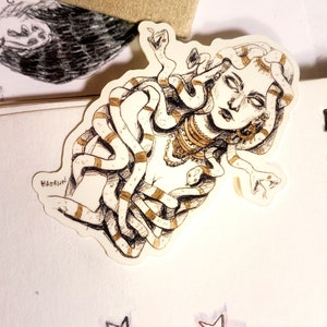 Op de afbeelding: Een sticker met een gedetailleerde illustratie van Medusa met slangen als haar. Het kunstwerk is in zwart en wit met gouden accenten, en de naam van de kunstenaar, "Hadrien", is zichtbaar. De sticker bevindt zich op een wit oppervlak met stervormige decoraties.