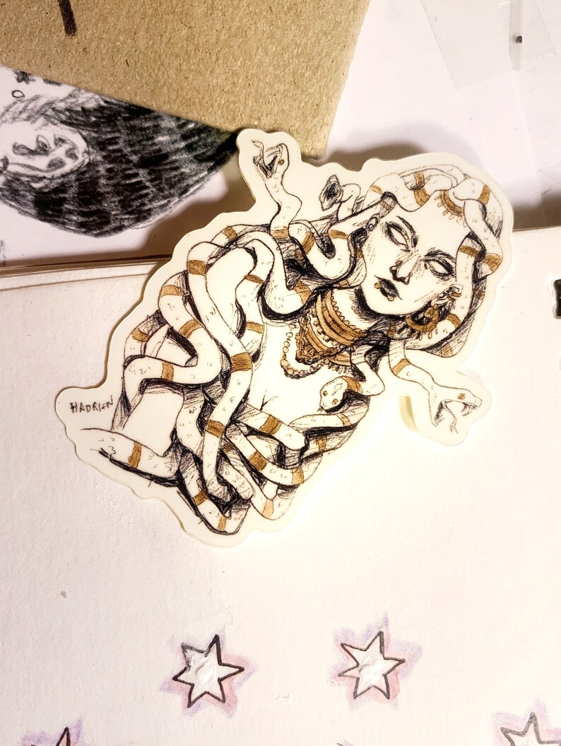 Op de afbeelding: Een sticker met een gedetailleerde illustratie van Medusa met slangen als haar. Het kunstwerk is in zwart en wit met gouden accenten, en de naam van de kunstenaar, "Hadrien", is zichtbaar. De sticker bevindt zich op een wit oppervlak met stervormige decoraties.