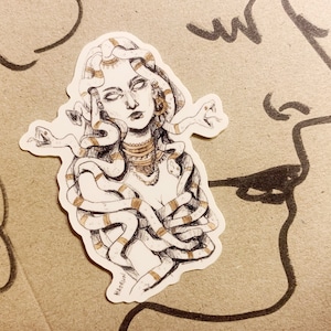 Op de afbeelding: Een sticker met een gedetailleerde illustratie van Medusa, met slangen als haar en sierlijke sieraden. Het kunstwerk is in zwart-wit met gouden accenten, op een neutrale achtergrond.