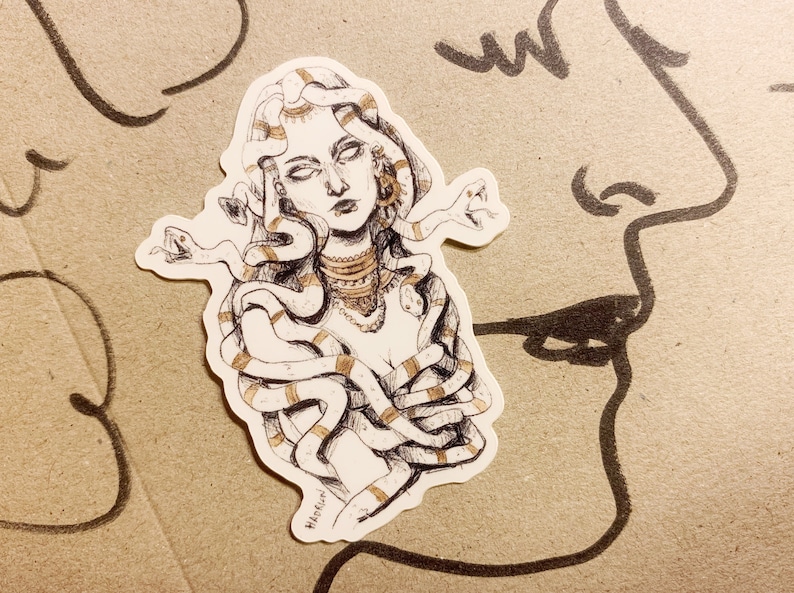 Op de afbeelding: Een sticker met een gedetailleerde illustratie van Medusa, met slangen als haar en sierlijke sieraden. Het kunstwerk is in zwart-wit met gouden accenten, op een neutrale achtergrond.