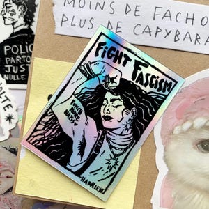 Puede incluir: Una pegatina hologr&aacute;fica con las palabras "FIGHT FASCISM" y una ilustraci&oacute;n de una persona con el pu&ntilde;o en alto. La pegatina tambi&eacute;n incluye el texto "PUNCH MORE NAZIS" y est&aacute; rodeada de otras pegatinas y texto escrito a mano.