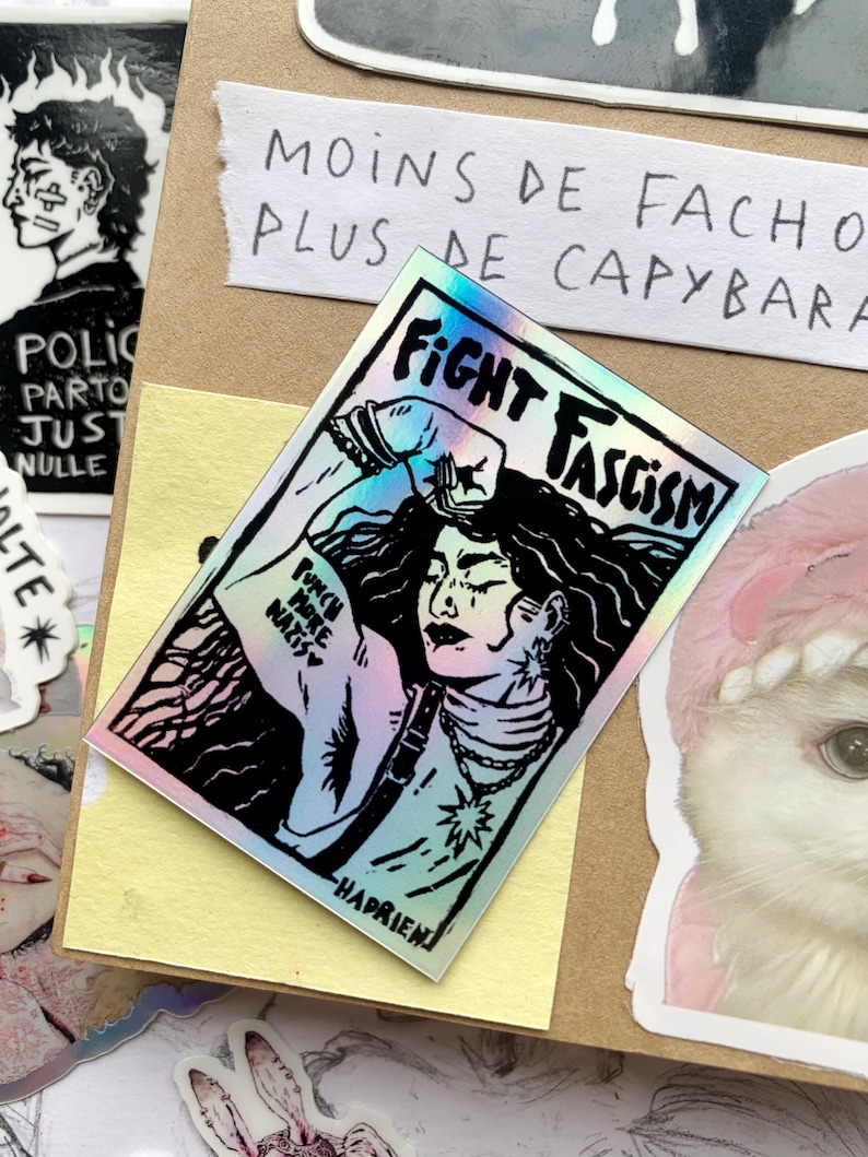 Puede incluir: Una pegatina hologr&aacute;fica con las palabras "FIGHT FASCISM" y una ilustraci&oacute;n de una persona con el pu&ntilde;o en alto. La pegatina tambi&eacute;n incluye el texto "PUNCH MORE NAZIS" y est&aacute; rodeada de otras pegatinas y texto escrito a mano.