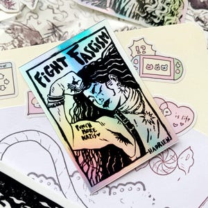 Puede incluir: Pegatina hologr&aacute;fica con las palabras "FIGHT FASCISM" y una ilustraci&oacute;n de una persona con el pu&ntilde;o en alto. La pegatina tambi&eacute;n incluye el texto "PUNCH MORE NAZIS" y la firma del artista. Otras pegatinas son visibles en el fondo.