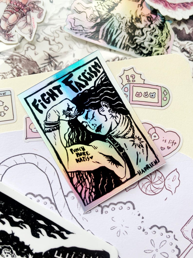 Puede incluir: Pegatina hologr&aacute;fica con las palabras "FIGHT FASCISM" y una ilustraci&oacute;n de una persona con el pu&ntilde;o en alto. La pegatina tambi&eacute;n incluye el texto "PUNCH MORE NAZIS" y la firma del artista. Otras pegatinas son visibles en el fondo.