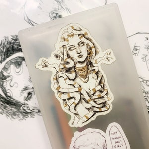 Op de afbeelding: Een sticker met een gedetailleerde illustratie van Medusa met slangen als haar, versierd met gouden accenten. De sticker is geplaatst op een doorschijnende container, met andere schetsen op de achtergrond. De sticker heeft een handgetekende uitstraling.