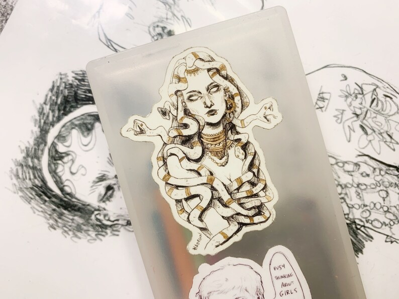 Op de afbeelding: Een sticker met een gedetailleerde illustratie van Medusa met slangen als haar, versierd met gouden accenten. De sticker is geplaatst op een doorschijnende container, met andere schetsen op de achtergrond. De sticker heeft een handgetekende uitstraling.
