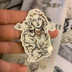 Op de afbeelding: Een sticker met een gedetailleerde illustratie van Medusa met slangen als haar. Het kunstwerk is in zwart en wit met gouden accenten. De handtekening van de kunstenaar is zichtbaar.