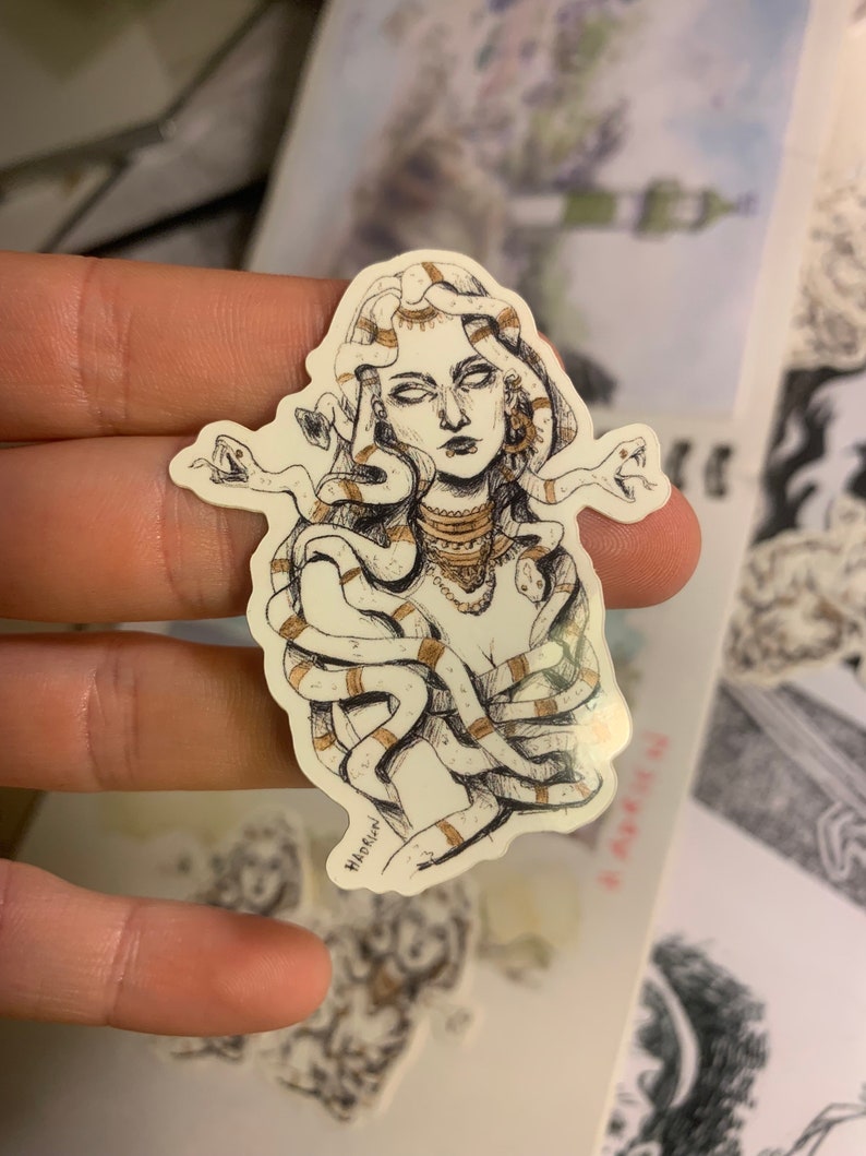 Op de afbeelding: Een sticker met een gedetailleerde illustratie van Medusa met slangen als haar. Het kunstwerk is in zwart en wit met gouden accenten. De handtekening van de kunstenaar is zichtbaar.