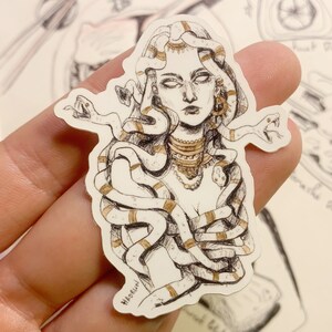 Op de afbeelding: Een sticker met een gedetailleerde illustratie van Medusa met slangen als haar, versierd met gouden accenten. Het kunstwerk is in zwart-wit met gouden highlights en toont een mythologisch thema.