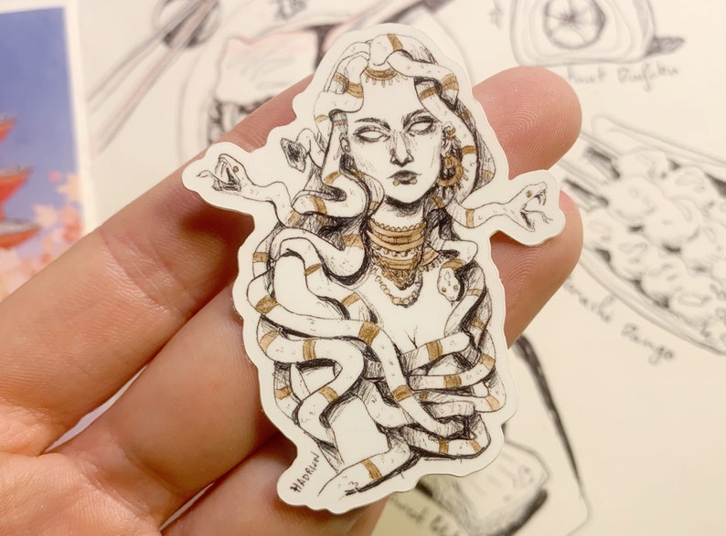 Op de afbeelding: Een sticker met een gedetailleerde illustratie van Medusa met slangen als haar, versierd met gouden accenten. Het kunstwerk is in zwart-wit met gouden highlights en toont een mythologisch thema.
