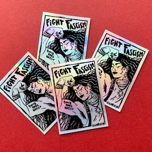 Puede incluir: Cuatro pegatinas hologr&aacute;ficas con el texto "FIGHT FASCISM" y una ilustraci&oacute;n de una persona con el pu&ntilde;o en alto. Las pegatinas tambi&eacute;n incluyen el texto "PUNCH MORE NAZIS" y la firma del artista.