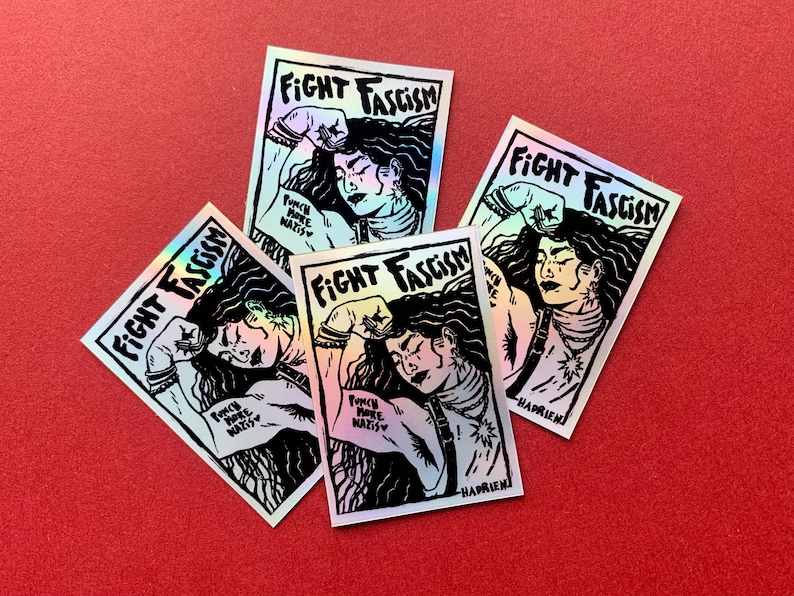 Puede incluir: Cuatro pegatinas hologr&aacute;ficas con el texto "FIGHT FASCISM" y una ilustraci&oacute;n de una persona con el pu&ntilde;o en alto. Las pegatinas tambi&eacute;n incluyen el texto "PUNCH MORE NAZIS" y la firma del artista.