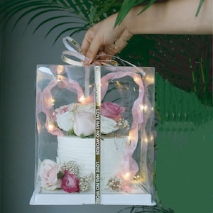 May include: A white cake decorated with pink and white flowers is displayed in a clear plastic box with a pink ribbon and string lights. The box is tied with a gold ribbon that says "LOVE HAS NO WORDS".