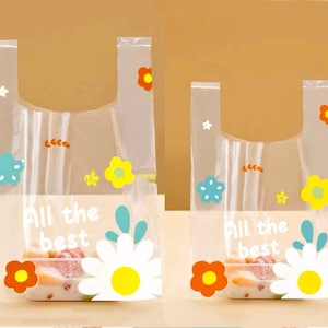 Puede incluir: Dos bolsas de pl&aacute;stico transparentes con asas, con un dise&ntilde;o floral colorido y el texto "All the best". Las bolsas est&aacute;n vac&iacute;as y listas para llenarse con regalos o golosinas.