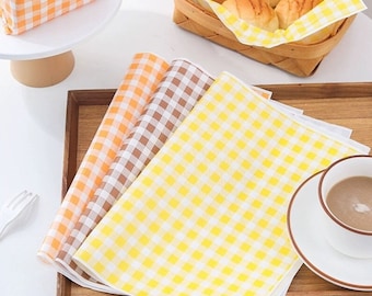 May include: Three checkered paper sheets in yellow, orange, and brown. The sheets are folded and stacked on a wooden tray. The sheets are perfect for wrapping sandwiches, snacks, or other food items.
