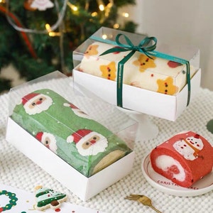 May include: Three clear plastic boxes with white lids containing Christmas themed decorated sponge cakes. The cakes are decorated with Santa Claus, reindeer, and snowman designs. The boxes are tied with green ribbon. The boxes are sitting on a white surface.