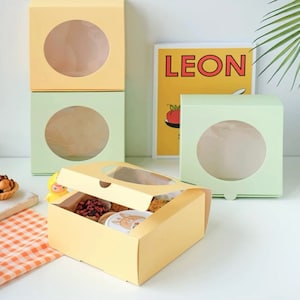 May include: Three yellow and green cardboard boxes with clear circular windows. The boxes are stacked on top of each other. The top box is open and has a variety of small pastries inside. The boxes are ideal for packaging and displaying small baked goods.