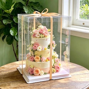 May include: A three-tiered cake with smooth cream-colored frosting, decorated with an assortment of pink, peach, and white roses and small yellow flowers. The cake is enclosed in a tall, clear, square plastic display box tied with a gold ribbon.