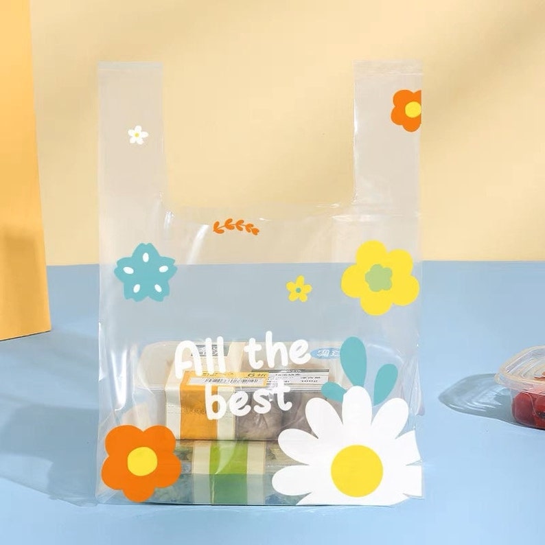 Puede incluir: Una bolsa de la compra reutilizable de pl&aacute;stico transparente con una margarita blanca, flores naranjas, amarillas y azules y hojas verdes impresas en ella. La bolsa tiene un asa y dice "All the best" en texto negro.
