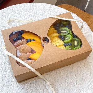 May include: A brown cardboard box with clear plastic windows shaped like a bow. The box is filled with sliced fruit, including mango, pineapple, kiwi, and blueberries.