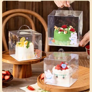 May include: Three clear plastic boxes with handles, each containing a small cake decorated with fruit and frosting. The boxes have cute cartoon stickers on them.