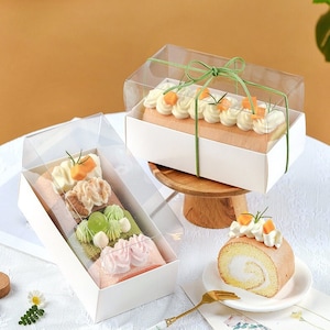 May include: Two clear plastic boxes with white lids containing different types of decorated cake rolls. The boxes are tied with green ribbon. A single cake roll is on a white plate with a gold fork.