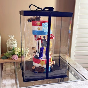 May include: A clear plastic box with a black lid and a black ribbon. Inside the box is a multi-tiered cake decorated with a Transformers character, a car, and stars. The cake is on a black base and has a gold star on top. The cake is surrounded by gold and silver decorations. The text "Happy Birthday" is written on the cake.