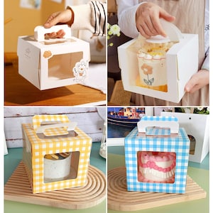 May include: Four cake boxes with clear windows and handles. Two boxes are white with cartoon designs, one is yellow with a checkered pattern, and one is blue with a checkered pattern.